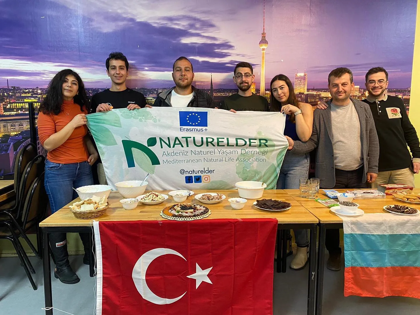 Participants holding a NATURELDER banner behind tables of food at the gathering in Berlin