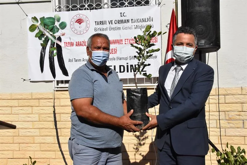 Proje afişinin önünde birlikte keçiboynuzu fidanı tutan iki kişi