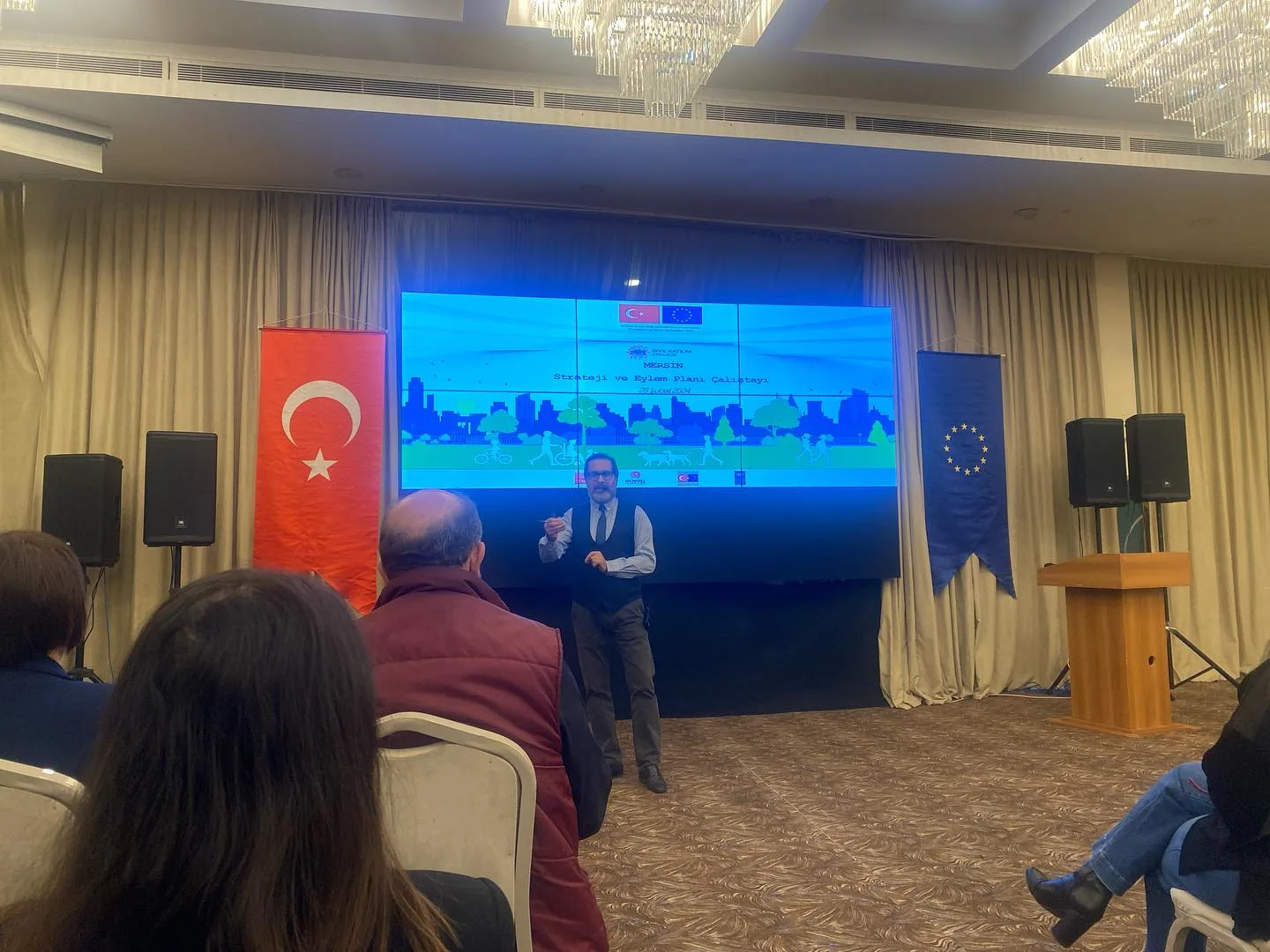 A speaker and seated participants in front of a screen displaying the Mersin Strategy and Action Plan Workshop title