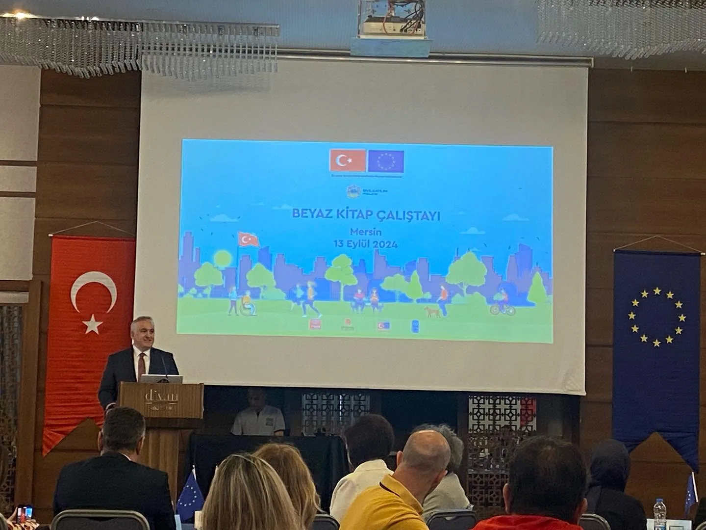 Mersin’de Beyaz Kitap Çalıştayı başlığı ve 13 Eylül 2024 tarihi bulunan sunum ekranının önünde konuşmacı ve salondaki katılımcılar